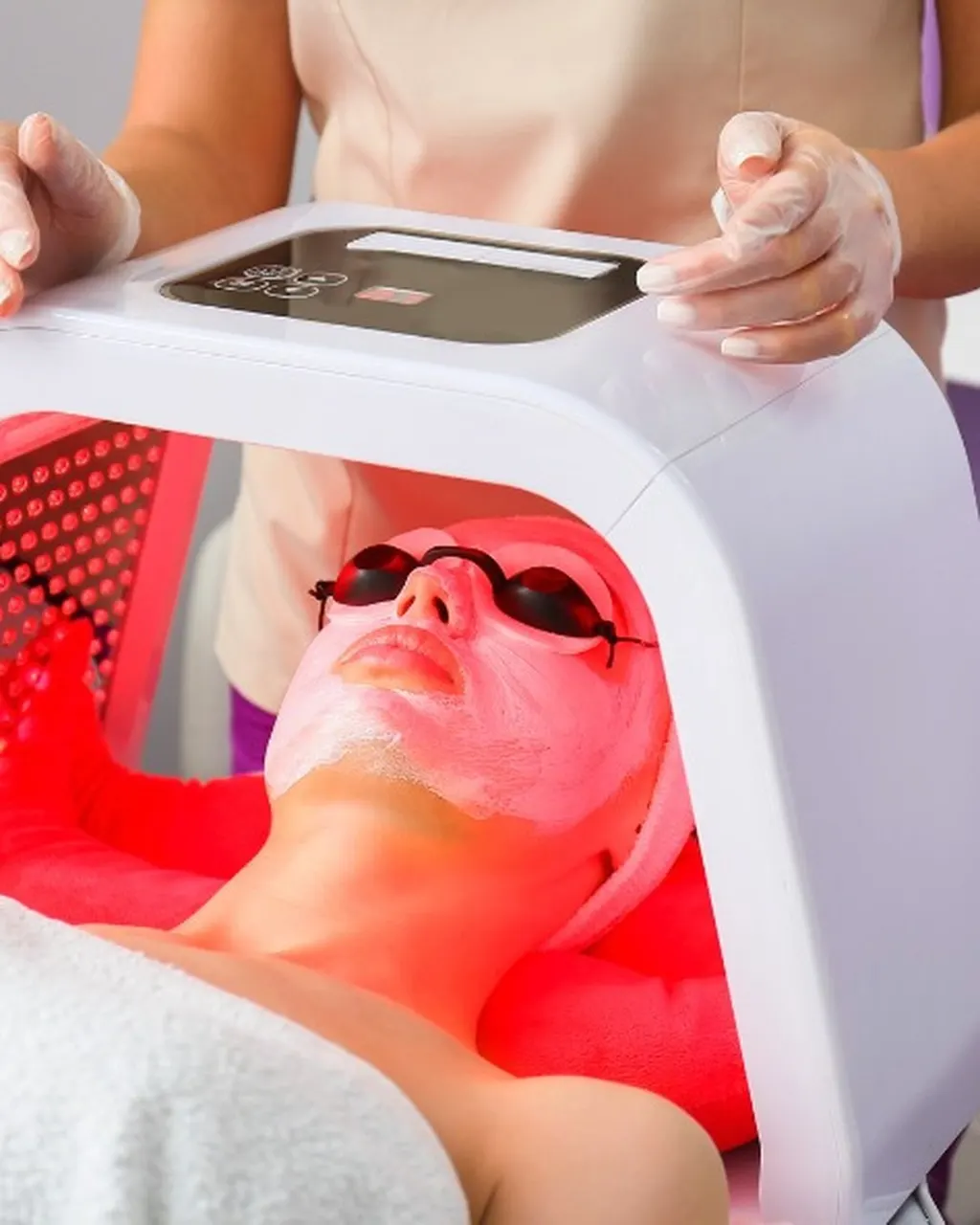 An esthetician in gloves positioning an LED light mask over a client during a facial